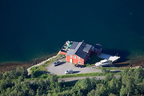 Rotes Fischerhaus in Nord-Norwegen, direkt am klaren, blauen Wasser. Ein Boot liegt an der Anlegestelle, umgeben von grüner Natur. Perfekter Ausgangspunkt für Angelabenteuer und Naturliebhaber. Ideal für Angelreisen und Entspannung.