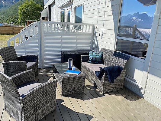 Gemütliche Terrasse in Nord-Norwegen: Moderne Loungemöbel aus Rattan auf Holzdeck mit atemberaubendem Blick auf schneebedeckte Berge am Fjord. Perfekter Ort zum Entspannen und Genießen der spektakulären nordischen Landschaft.