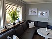 Gemütliches Wohnzimmer mit grauem Ecksofa, rundem Tisch, großem Fenster mit Blick auf grüne Pflanzen und zwei gerahmten Bildern an der Wand, darunter eines mit erkennbaren Fischarten.