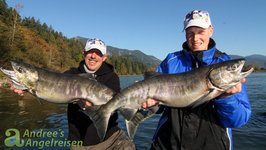 Zwei Angler halten lachend große Lachse vor einer malerischen Flusslandschaft in die Kamera.