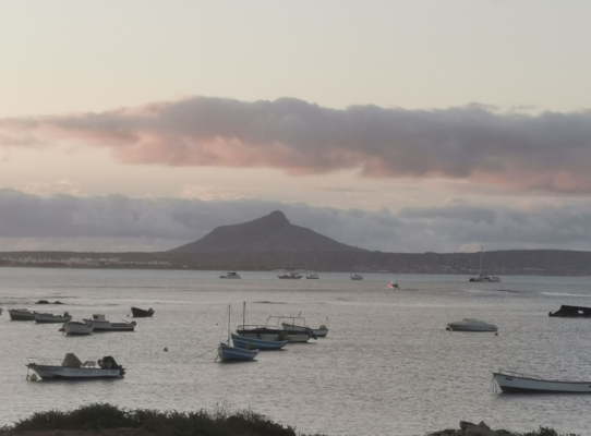 Sonnenuntergang über der Bucht von São Vicente, Kapverden: Boote schwimmen im ruhigen Meer, während der Himmel in sanften Rosa- und Grautönen leuchtet. Im Hintergrund erhebt sich ein beeindruckender Berg. Serene Meereslandschaft.