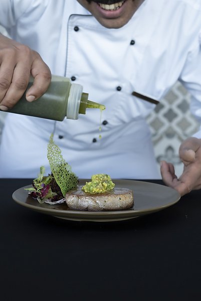 Ein Koch in Süd-Norwegen verfeinert kunstvoll ein Gourmetgericht mit Sauce, bestehend aus gegrilltem Fisch, knackigem Salat und einer grünen Dekoration auf einem Teller. Kulinarische Erlebnisse und regionale Küche genießen.