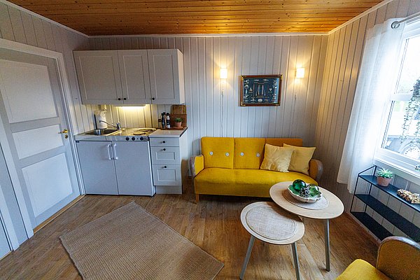Gemütliches Ferienhaus in Hitra, Norwegen: heller Raum mit gelbem Sofa, rustikaler Holzwand, kleiner Küchenzeile und skandinavischem Dekor. Ideal für Angelurlaub und Erholung in malerischer Umgebung. Perfekte Ausgangsbasis für Abenteuer in der Natur.
