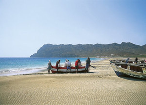 Eine Gruppe von Menschen steht an einem sandigen Strand neben einem kleinen Boot, während im Hintergrund das Meer und eine felsige Küstenlandschaft sichtbar sind. Eine Gruppe von Menschen steht an einem sandigen Strand neben einem kleinen Boot, während im Hintergrund das Meer und eine felsige Küstenlandschaft sichtbar sind.