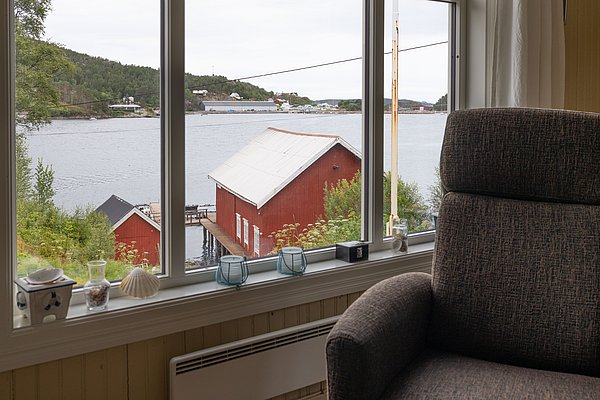 Blick aus einem gemütlichen Zimmer auf ein rotes Bootshaus am Wasser in Norwegen. Malerische Landschaft der Region Hitra, ideal für einen erholsamen Angelurlaub. Naturnahe Entspannung am malerischen Fjord.