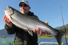Ein Angler präsentiert stolz einen frisch gefangenen Lachs auf einem Boot, umgeben von malerischer Landschaft und blauem Himmel.