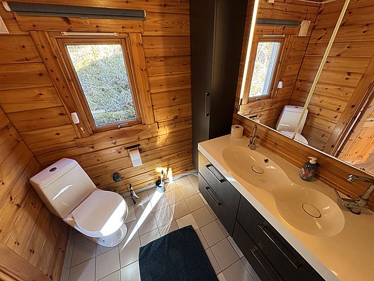 Modernes Badezimmer in einer Holzhütte auf Hitra, Norwegen. Helle Holzwände, Doppelwaschbecken, zwei Fenster, Toilette. Ideal für Anglerurlaub, kombiniert Komfort mit Naturerlebnis in skandinavischem Design. Entdecke Angelreisen in wunderschöner Umgebung.