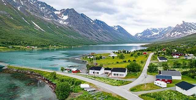 Luftaufnahme einer idyllischen Fjordlandschaft in Nord-Norwegen: klare Gewässer, malerische Hütten, majestätische Berge mit Schnee. Perfektes Reiseziel für Angelurlaub und Naturerlebnisse in atemberaubender skandinavischer Kulisse.