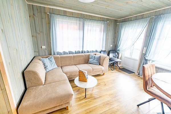 Gemütlicher Wohnbereich in Nord-Norwegen mit beiger Eckcouch, kleinem Tisch, Holzfußboden und großen Fenstern. Ideal für entspannte Angelreisen. Helles, einladendes Ambiente mit skandinavischem Flair. Perfekter Rückzugsort nach einem gelungenen Angeltag.
