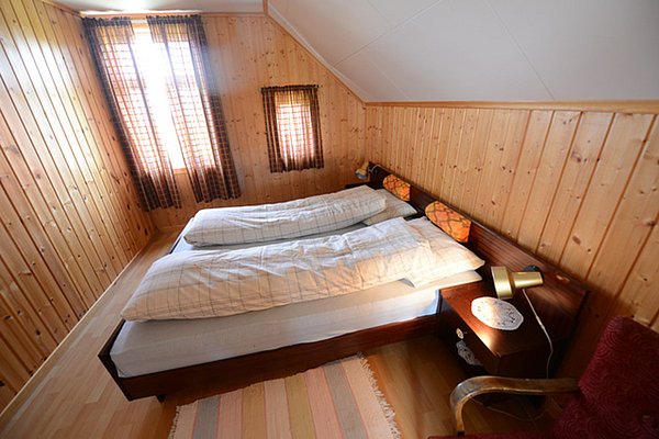 Gemütliches Schlafzimmer in Nordland, Norwegen: Holzwände, Doppelbett, Fenster mit Vorhängen, Nachttischlampe und Teppich. Ideal für erholsame Nächte nach einem Angeltag. Perfekte Unterkunft für Angelreisen in Norwegens idyllischer Natur.