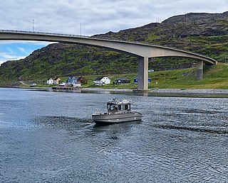 Ein kleines Motorboot fährt unter einer Brücke auf einem Fluss entlang, während im Hintergrund mehrere bunte Häuser am grünen Hang stehen.