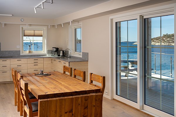 Moderne Küche mit Holztisch und Meerblick in Hitra, Norwegen. Große Fenster bieten Blick auf das klare Wasser und die malerische Küstenlandschaft. Ideal für entspannte Angelurlaube und kulinarische Erlebnisse.