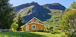 Gelbes Holzhaus vor majestätischer Bergkulisse in Nordland, Norwegen. Üppiges Grün umgibt das Haus unter sonnigem Himmel. Perfekter Ausgangspunkt für Angelurlaub und Naturerlebnisse.
