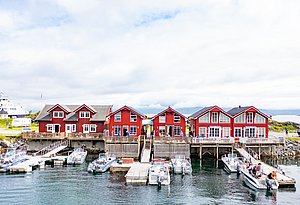 Rote Fischerhütten im typischen skandinavischen Stil an Norwegens Küste, umgeben von blauen Booten am Steg. Diese idyllische Szene in Nordland lädt zum Angelurlaub und Entdecken der malerischen Natur Norwegens ein.