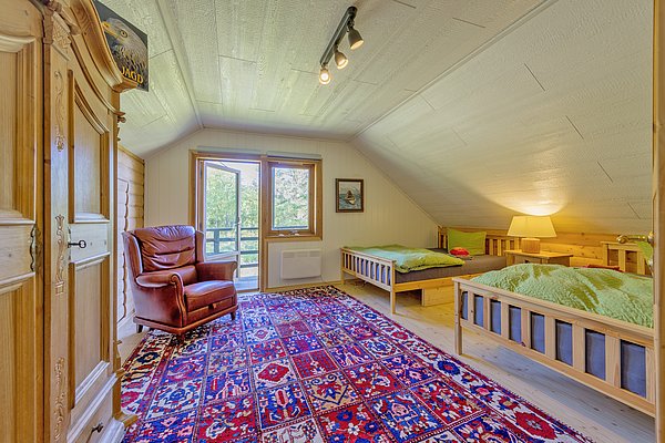 Gemütliches Schlafzimmer in einem norwegischen Holzhaus in Nordland, Norwegen. Rustikale Einrichtung mit Betten, rotem Sessel und farbigem Teppich. Fensterblick auf grüne Natur. Perfekt für einen erholsamen Angelurlaub. Ideal für Natur- und Norwegenliebhaber.