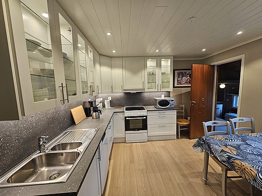 Moderne Küche in Südwest-Norwegen: Helle Schränke, Holzdecke, Edelstahlspüle, Geschirrspüler, Herd, Mikrowelle und Esstisch mit bunter Tischdecke. Ideal für Angelurlauber, die Komfort und Selbstversorgermöglichkeiten in gemütlicher Umgebung suchen.