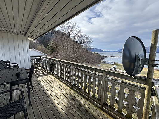 Überdachte Terrasse mit Holzboden, Blick auf norwegische Fjordlandschaft. Sitzmöglichkeiten und Satellitenschüssel links im Bild, ruhige Natur mit Bergen, Meer und Wolken im Hintergrund. Ideal für Angelurlaub in Hitra, Norwegen.