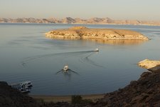 Ein weites Gewässer mit zwei Booten, die Spuren auf der Wasseroberfläche hinterlassen, und einer Insel in der Mitte, umgeben von einer kargen, bergigen Landschaft.