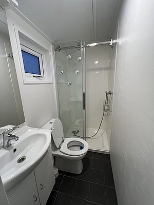 Modernes Badezimmer in Nord-Norwegen mit Dusche, weißer Wand, Waschbecken und Toilette. Helle Einrichtung und grauer Boden schaffen eine saubere, einladende Atmosphäre. Ideal für Reisende bei einem Angelurlaub in der Region.