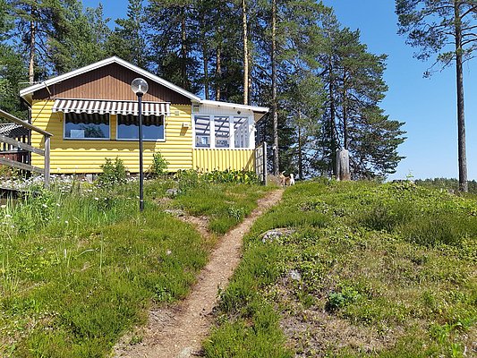 Ein gelbes Ferienhaus mit Markise steht inmitten einer grasbewachsenen und mit Kiefern bewachsenen Landschaft unter einem strahlend blauen Himmel.