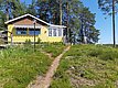 Ein gelbes Ferienhaus mit Markise steht inmitten einer grasbewachsenen und mit Kiefern bewachsenen Landschaft unter einem strahlend blauen Himmel.
