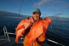 Ein Angler auf einem Boot hält zwei große, rote Fische vor dem blauen Himmel und Meer. Ein Angler auf einem Boot hält zwei große, rote Fische vor dem blauen Himmel und Meer.