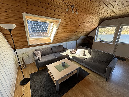 Gemütlicher Wohnraum in Hitra, Norwegen, mit Dachschräge, grauen Sofas und Blick aufs Wasser. Perfekt für Angelurlauber, die Komfort und Naturerlebnis kombinieren möchten. Ideal für entspannte Abende nach erfolgreichen Angelausflügen.