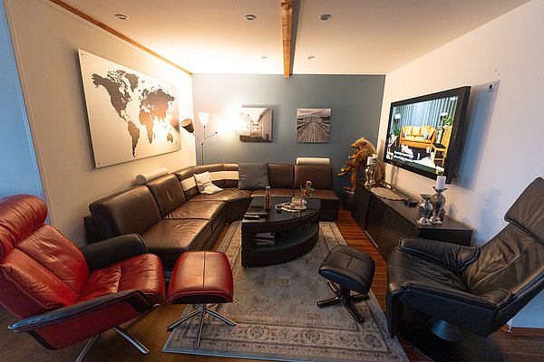 Gemütliches Wohnzimmer in Nord-Norwegen mit Ledersofas, stilvollen Sesseln und moderner Dekoration. Ein großer Fernseher und eine Weltkarte verleihen dem Raum behaglichen Charme und laden zum Entspannen ein.