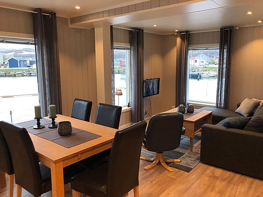 Gemütlicher Wohnbereich in Nord-Norwegen mit Holztisch, Ledersesseln und Blick aufs Wasser. Große Fenster bieten atemberaubende Aussicht, ideal für Angelurlaub und Entspannung nach einem abenteuerlichen Tag am Fjord. Perfekte Balance aus Komfort und Natur.