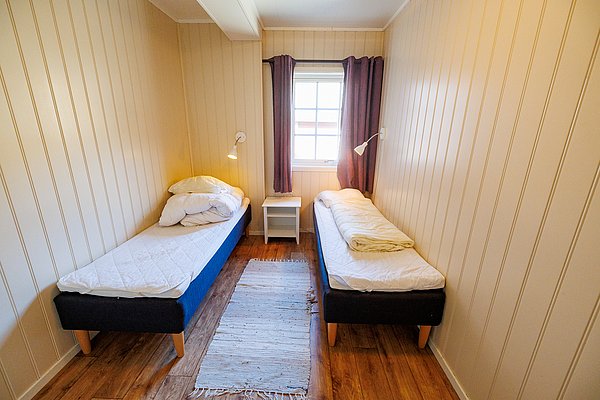 Gemütliches Schlafzimmer in Nordland, Norwegen, mit zwei Einzelbetten, Holzvertäfelung und Fenster. Perfekt für Angelurlaube, bietet Wärme und Erholung nach einem erlebnisreichen Tag am Wasser. Petri Heil – sorglos angeln weltweit!