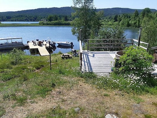 Am Ufer eines Sees befinden sich ein hölzerner Steg mit mehreren Booten, eine Terrasse und eine grüne, blühende Wiese mit Blick auf einen bewaldeten Horizont.
