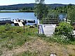 Am Ufer eines Sees befinden sich ein hölzerner Steg mit mehreren Booten, eine Terrasse und eine grüne, blühende Wiese mit Blick auf einen bewaldeten Horizont.