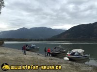 Drei Boote liegen am Ufer eines Sees vor einer Berglandschaft, während mehrere Personen sich am Ufer unterhalten; die Website "stefan-seuss.de" ist in der Ecke zu sehen.