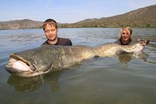 Zwei Personen posieren im Wasser mit einem riesigen Wels, umgeben von einem See und hügeliger Landschaft im Hintergrund.