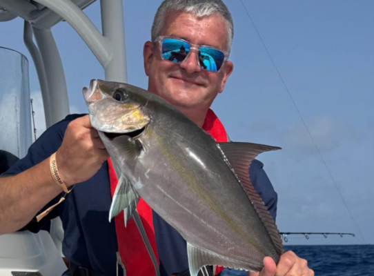 Ein Angler hält stolz einen gefangenen Fisch auf einem Boot. Der blaue Ozean und der Himmel von Sao Vicente, Kapverden, sind im Hintergrund zu sehen. Perfekter Spot für ein aufregendes Angelabenteuer.