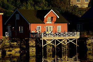 In der Abendsonne erstrahlt ein rotes Holzhaus auf Stelzen am Wasser in Hitra, Norwegen. Perfekt für Angelreisen, bietet es idyllische Ruhe und direkten Zugang zum fischreichen Meer.