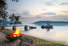 Ein idyllisches Küstenerlebnis in Südwest-Norwegen: Ein Boot schwimmt friedlich im ruhigen Wasser, während ein gemütliches Lagerfeuer am Ufer Wärme spendet. Perfekt für entspannte Angelreisen und Outdoor-Abenteuer.
