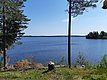 Ein malerischer See mit klarem, blauem Wasser, umgeben von grünen Bäumen und einem Gartenstuhl mit einer Tasse darauf im Vordergrund.