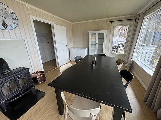 Gemütlicher Essbereich in einer Ferienunterkunft in Hitra, Norwegen. Helle Holzböden, Kaminofen, großer Esstisch mit Stühlen, Vitrine und viel Tageslicht durch große Fenster, die den Blick auf die Umgebung bieten.