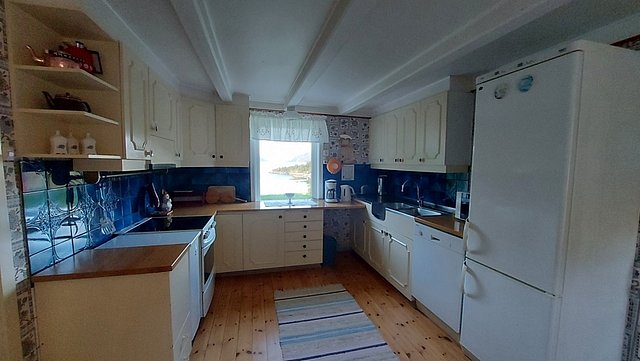 Gemütliche Küche in Nordland, Norwegen. Weiße Schränke, blauer Fliesenspiegel und großer Kühlschrank. Helles Fenster mit Blick in die Natur. Idealer Ausgangspunkt für Angelreisen und skandinavische Kochabenteuer. Authentisches, einladendes Ambiente.