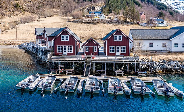 Rote Holzhäuser auf Stelzen am Wasser in Nordland, Norwegen. Davor Boote an einem Steg. Malerische Landschaft mit Hügeln und ländlicher Idylle. Perfekt für Angelreisen und Entspannung am Fjord.