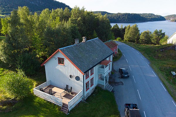 Luftaufnahme eines traditionellen roten Hauses mit Terrasse in Hitra, Norwegen. Umgeben von grüner Landschaft und in der Nähe eines malerischen Sees. Perfekte Kulisse für Angelreisen und Naturerlebnisse in Skandinavien.