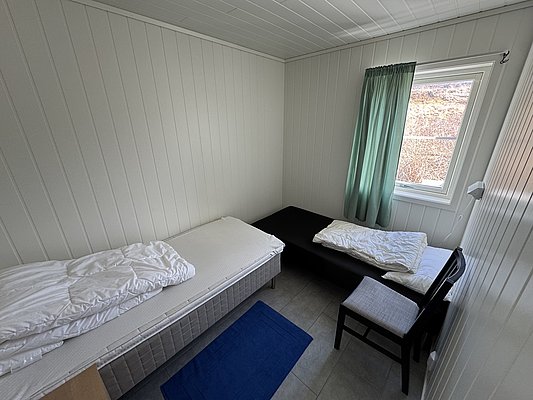 Einfache, gemütliche Unterkunft in der Region Hitra, Norwegen. Zwei Betten mit schlichter weißer Bettwäsche, ein Fenster mit grünen Vorhängen und ein blauer Teppich schaffen eine entspannte Atmosphäre. Ideal für Angelurlauber.