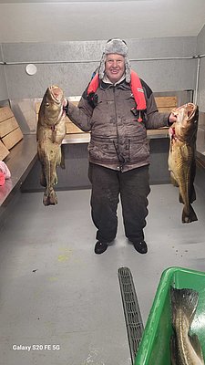 Ein Angler in Nordland, Norwegen, hält stolz zwei große Dorsche. Er trägt warme Kleidung und eine orangefarbene Rettungsweste. Perfekte Bedingungen für unvergessliche Angelreisen in Skandinavien.