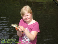 Ein Kind in einem pinken T-Shirt hält lächelnd einen Fisch, vermutlich einen Flussbarsch, vor einem Gewässer.