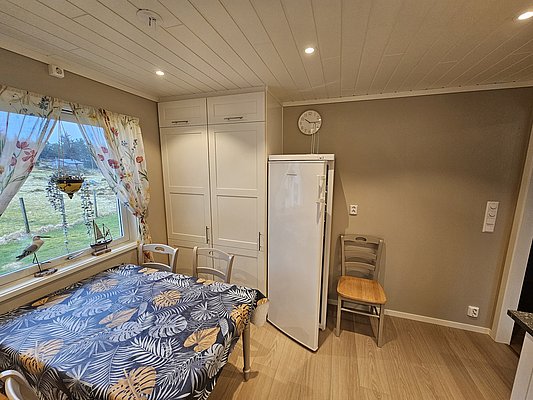 Gemütliche Küche in Südwest-Norwegen mit Tisch, buntem Tischtuch und Fensterblick ins Grüne. Helle Einrichtung, Kühlschrank und dekorative Vorhänge schaffen entspannte Atmosphäre. Ideal für Angelurlaub und Erholung.