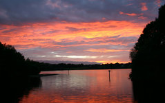 Ein ruhiger See bei Sonnenuntergang mit einem dramatisch rot-orange gefärbten Himmel spiegelt sich im Wasser wider, umgeben von Bäumen.