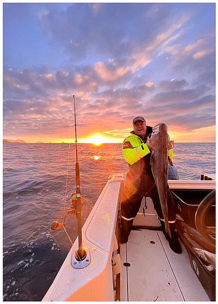 Eine Person in leuchtender Kleidung hält einen großen Fisch an Bord eines Bootes bei Sonnenuntergang auf dem Meer.