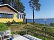 Eine gelbe Holzhütte mit gestreifter Markise steht an einem sonnigen Seeufer mit blühenden Gänseblümchen und Kiefern im Hintergrund.