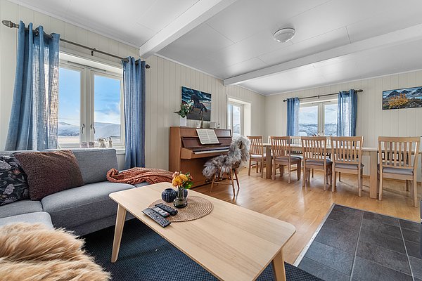 Gemütliches Wohnzimmer in Nord-Norwegen mit hellen Holzmöbeln, Klavier, großem Esstisch und Panoramablick auf verschneite Berge durch große Fenster. Perfekt für einen erholsamen Angelurlaub in skandinavischer Atmosphäre.
